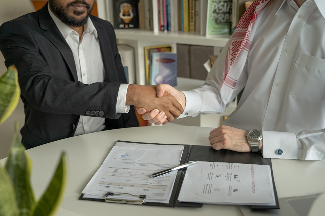 Two professionals in business attire shaking hands over a desk with a real estate contract and a pen, signifying a successful property sale agreement.