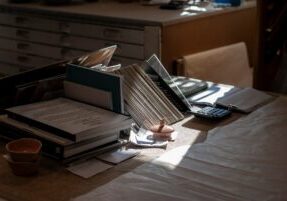 Homeowner sits at a sunlit dining table with a calculator, pen, and a stack of papers, looking concerned as they calculate...